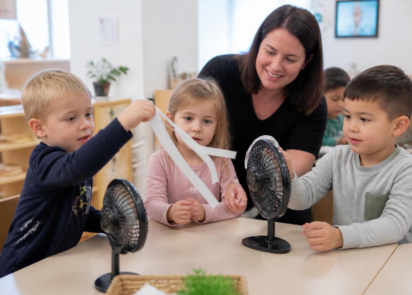 Preschool children experimenting with airflow using lightweight paper strips and small fans at a classroom table